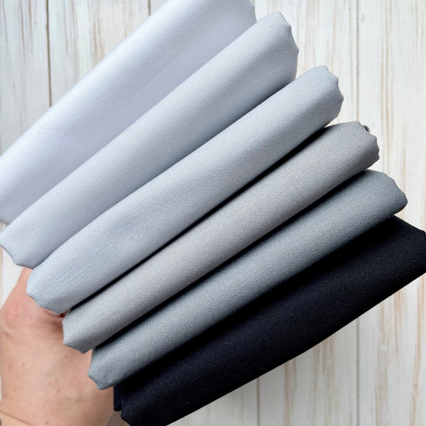 Six folded half metre cuts of cotton fabric ranging from white to black with four shades of grey.